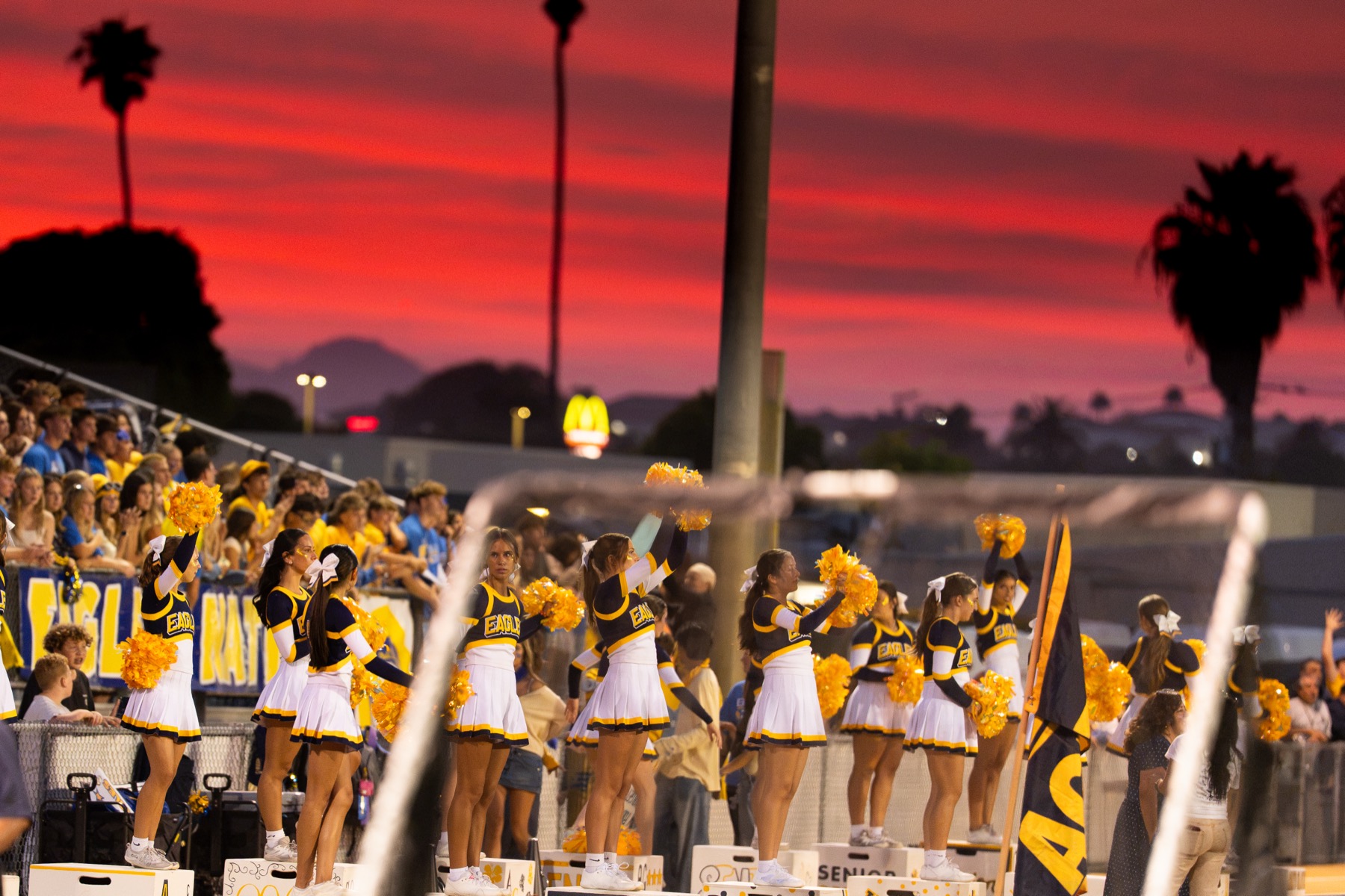 Arroyo Grande cheerleaders raising their pom-poms in front of a deep red sunset sky